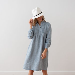May include: A woman wearing a light blue linen dress with a white hat. The dress has a button-up front and a pleated design.