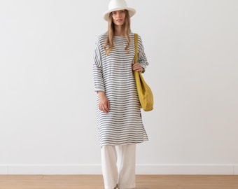May include: A woman in a white hat, a long striped dress, white trousers, and sandals, carrying a yellow shoulder bag. The dress features navy blue horizontal stripes. The outfit is casual and suitable for warmer weather.