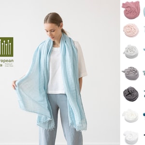 Peut inclure: Une écharpe en lin bleu clair avec des bords effilochés. L'écharpe est faite de lin européen, une fibre de lin de qualité supérieure. L'écharpe est drapé autour du cou d'une personne portant un t-shirt blanc et un pantalon gris.