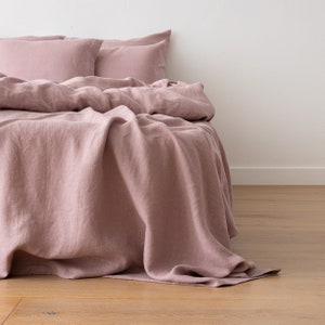 May include: A dusty rose linen bedding set with a flat sheet, fitted sheet, and two pillowcases. The bedding is arranged on a bed with the sheet draped over the side.