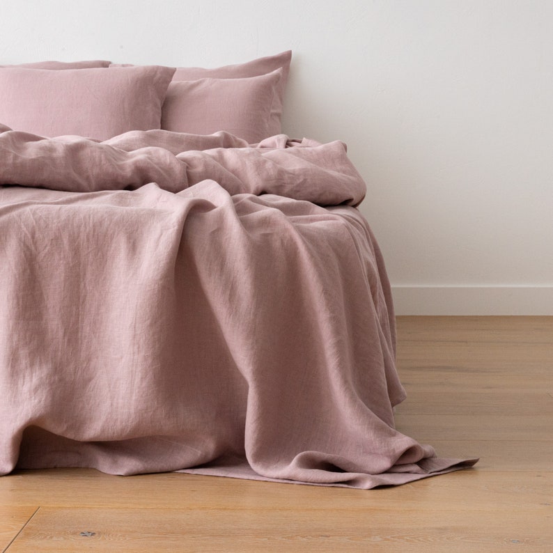 May include: A dusty rose linen bedding set with a flat sheet, fitted sheet, and two pillowcases. The bedding is arranged on a bed with the sheet draped over the side.