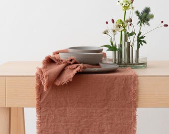 May include: A terracotta-colored linen table runner with frayed edges, topped with a stack of two gray ceramic bowls on a matching plate, and a clear glass multi-stem flower vase containing various green and white floral stems.