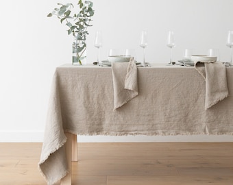 May include: A light brown linen tablecloth with frayed edges, set on a wooden table. The tablecloth is decorated with four place settings, each with a white ceramic plate, a linen napkin, and a clear glass. A vase with green eucalyptus branches sits in the center of the table.