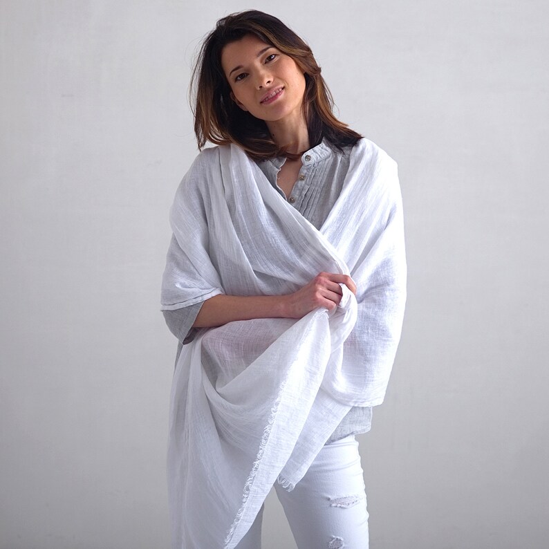 May include: A white linen scarf draped around a person's shoulders. The scarf is lightweight and has a soft, flowing texture.