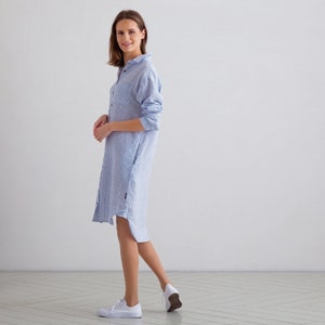 Puede incluir: Un vestido camisa de lino azul claro con botones en la parte delantera y mangas largas. El vestido tiene un corte holgado y una silueta ligeramente en A.
