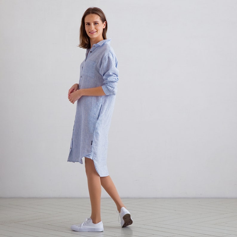 Puede incluir: Un vestido camisa de lino azul claro con botones en la parte delantera y mangas largas. El vestido tiene un corte holgado y una silueta ligeramente en A.