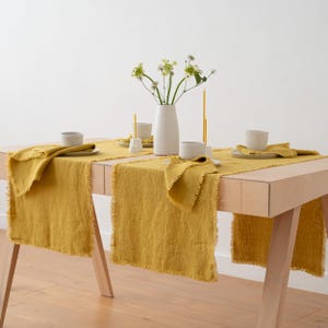 Puede incluir: Una mesa de comedor con un camino de mesa de lino amarillo mostaza y servilletas a juego. La mesa está puesta con tazas, platos y un jarrón blanco con flores. La mesa tiene un acabado de madera clara.