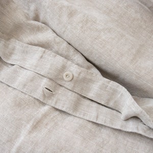 May include: Close-up of a beige linen fabric with a white button. The fabric has a subtle texture and a slightly wrinkled appearance.