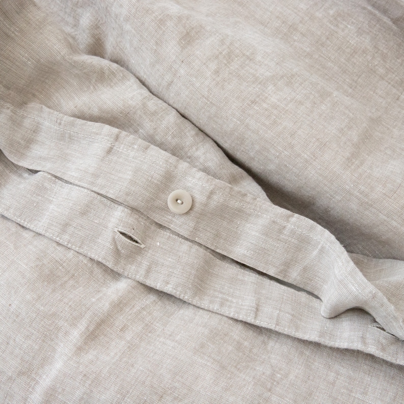 May include: Close-up of a beige linen fabric with a white button. The fabric has a subtle texture and a slightly wrinkled appearance.