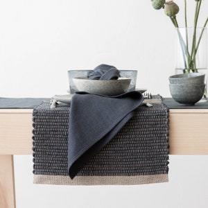 May include: A dark gray woven placemat with a light brown border. A dark gray linen napkin is folded on top of the placemat. The placemat is on a light wood table.