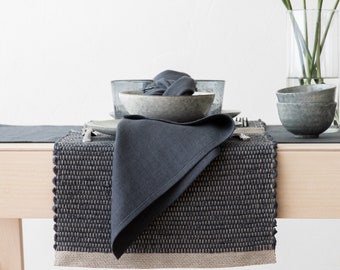 May include: A dark gray woven placemat with a light brown border. A dark gray linen napkin is folded on top of the placemat. The placemat is on a light wood table.