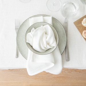 Puede incluir: Una mesa formal con un mantel de lino blanco. Un plato gris contiene una servilleta blanca y un cuenco pequeño con una servilleta blanca doblada. Un tenedor y un cuchillo están a ambos lados del plato. También hay un vaso de agua y una tabla de madera con comida.