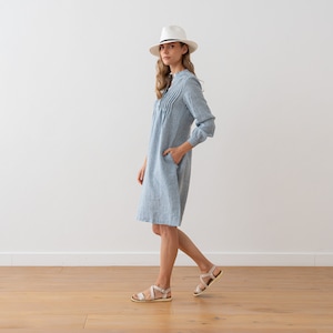 May include: A woman wearing a light blue linen dress with a white hat and sandals. The dress has a gathered neckline and long sleeves.