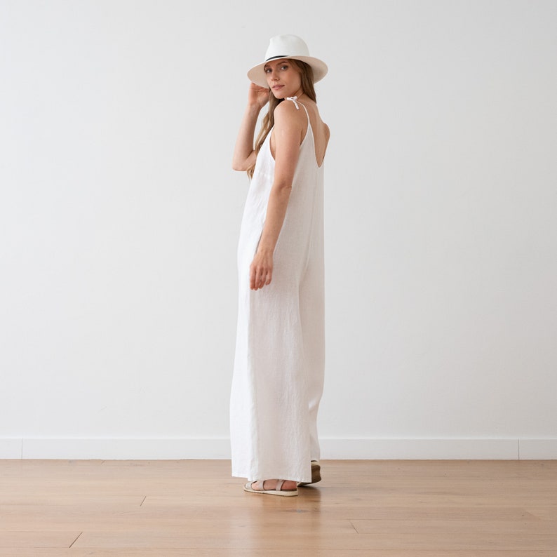May include: A white linen jumpsuit with thin straps and a wide leg. The model is wearing a white hat and sandals.