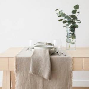 May include: A light brown linen table runner with frayed edges, placed on a light wood table. Two ceramic bowls, two glasses, and two silver forks are set on the table runner. A vase with eucalyptus branches is on the table.