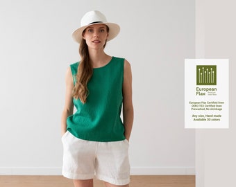 May include: A woman wearing a green sleeveless linen top and white linen shorts. She is wearing a white hat. The text on the sign reads: "European Flax. European Flax Certified linen Oeko Tex Certified linen Prewashed, No shrinkage Any size, Hand made Available 30 colours"