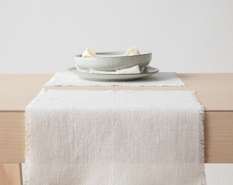 Puede incluir: Una mesa de comedor de tonos neutros con un camino de mesa de lino color crema con borde deshilachado. Un mantel individual a juego, platos y un cuenco con servilletas dobladas están dispuestos sobre la mesa, creando una estética minimalista.