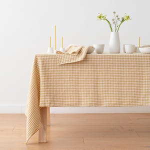 May include: A tan and white gingham tablecloth on a wooden table. White ceramic bowls, a vase with flowers, and yellow candles are arranged on the table. A matching gingham napkin is folded on the table.