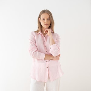 Pode incluir: Uma mulher vestindo uma camisa de linho rosa de manga comprida e botões. Ela está em pé em frente a um fundo branco.