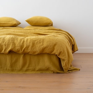 May include: A bed with mustard-yellow linen bedding, including a fitted sheet, duvet cover, and two pillowcases. The bedding is draped over a bed frame, with the fabric cascading to the floor. The room has a white wall and a wooden floor.