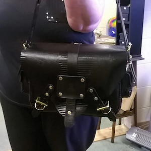 May include: A black leather satchel bag with a textured finish. The bag has a flap closure with two buckles and a strap with a metal buckle.
