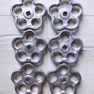 Może przedstawiać: Sześć srebrnych metalowych gałek do zaworów, każda z sześcioma otworami i w kształcie kwiatu z pięcioma płatkami. Każda gałka ma kwadrat w środku.