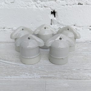 Könnte beinhalten: Eine Ansammlung von fünf hellgrauen Keramikobjekten, jedes mit einem abgerundeten Sockel und einer stilisierten, blütenblattförmigen Oberseite. Die Objekte sind eng beieinander angeordnet, mit einem kleinen Loch oben. Der Hintergrund ist eine weiße Backsteinmauer.