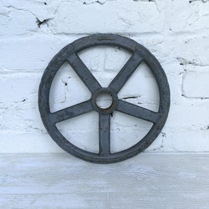 May include: A circular gray metal industrial wheel featuring five thick spokes radiating from a central hub. The surface of the wheel shows signs of wear with visible patches of rust and chipped paint, giving it a weathered, aged appearance.