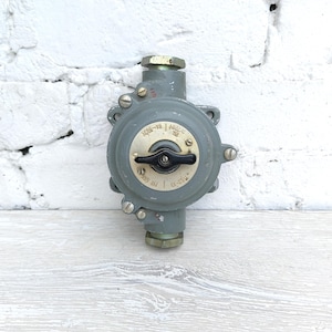 May include: A grey industrial electrical switch with a black lever and circular dial. The switch is mounted on a white brick wall and a white wooden surface. The dial has the numbers "4055-99" and "4022-70" printed on it.