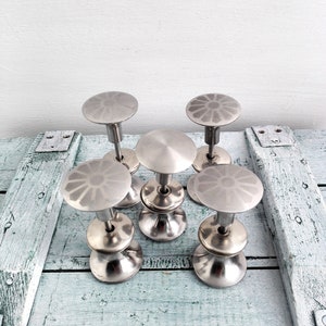 May include: Five silver metal cabinet knobs with a decorative flower design on the top. The knobs are arranged in a circle on a light blue wooden surface.