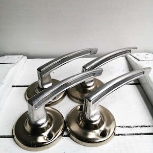 Op de afbeelding: Vier zilveren deurgrepen met een geborstelde afwerking en een gebogen ontwerp. De grepen zijn gemonteerd op ronde, goudkleurige bases.