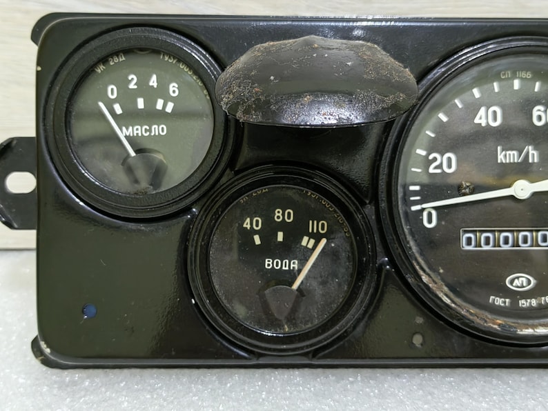 May include: A black dashboard with three gauges. The gauges are labeled "MASLO", "BODA", and "km/h". The "MASLO" gauge has a needle pointing to 4, the "BODA" gauge has a needle pointing to 80, and the "km/h" gauge has a needle pointing to 0. The "km/h" gauge also has a digital display that reads 00000.