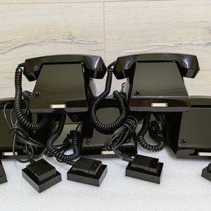 Può includere: Una pila di cinque telefoni rotativi neri con cavi a spirale. I telefoni sono disposti a forma di piramide, con il telefono superiore rivolto in avanti. I telefoni sono su una superficie bianca.