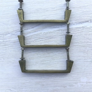 Peut inclure: Trois poignées de tiroir en métal couleur laiton avec une barre rectangulaire et des extrémités effilées. Chaque poignée comporte deux boulons filetés verticaux avec des rondelles fixées au sommet, s'étendant vers le haut depuis les extrémités.