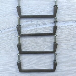 Peut inclure: Quatre poignées de tiroir rectangulaires en métal avec une finition bronze sombre et patinée. Chaque poignée présente deux montants verticaux avec des vis et des rondelles métalliques argentées fixées vers le haut du cadre.