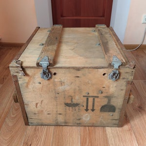May include: A wooden crate with two metal clasps and a handle on each side. The crate has a faded black and white symbol on the front. The symbol includes a drop of water, two arrows pointing down, and a flask.