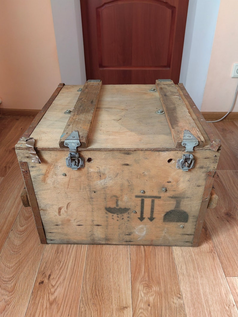 May include: A wooden crate with two metal clasps and a handle on each side. The crate has a faded black and white symbol on the front. The symbol includes a drop of water, two arrows pointing down, and a flask.