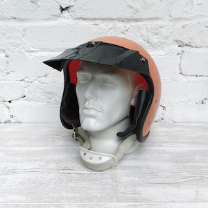 May include: An orange open-face motorcycle helmet featuring a detachable black visor, black interior padding, and a light grey chin strap with a white plastic buckle. The helmet is displayed on a white mannequin head to show its structure.
