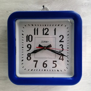 Puede incluir: Un reloj de pared cuadrado azul con una cara blanca y números negros. El reloj tiene una aguja de segundos roja y agujas de horas y minutos negras. La esfera del reloj también tiene la palabra "STŘEDA" impresa en ella.