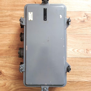 Può includere: Una scatola elettrica in metallo grigio con più connettori e un'etichetta sul davanti. La scatola è montata su una superficie in legno.