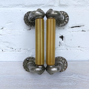May include: Two antique door handles. Each handle has a silver-coloured, ornate rosette and a curved top. The central section is a cylindrical, golden-yellow colour. The handles are mounted on a white brick wall.