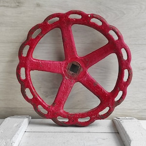 Könnte beinhalten: Ein großes, rotes Metallrad mit einem fünfspeichigen Design und einem quadratischen Loch in der Mitte. Das Rad hat einen dekorativen, gewellten Rand.