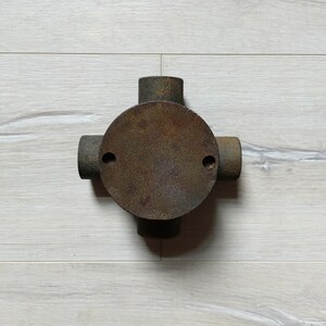 May include: A rusty metal junction box with four openings for electrical wiring. The box is round with a flat top and a slightly raised rim.