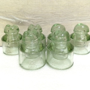 Puede incluir: Seis aisladores de vidrio transparente con un tono verde. Los aisladores tienen una parte superior redondeada y una base ancha. Los aisladores están marcados con el número "297".