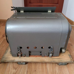 May include: A gray metal box with a wooden base and several latches. The box has a flat top and a slightly rounded bottom. The box is sitting on a wooden floor.