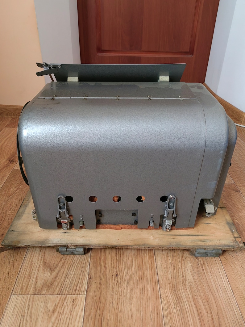 May include: A gray metal box with a wooden base and several latches. The box has a flat top and a slightly rounded bottom. The box is sitting on a wooden floor.