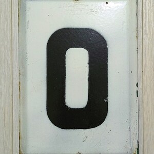 May include: Rectangular white metal sign with the number "0" in black. The sign has a distressed appearance with rust around the edges and two holes at the top for mounting. The background is a light wood grain.