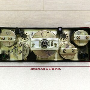 May include: A black metal panel with four round gauges, each with a silver face and black markings. The panel measures 12 3/16 inches wide and 4 5/8 inches tall.