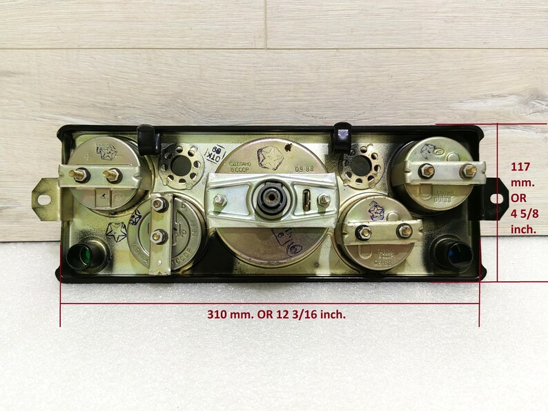 May include: A black metal panel with four round gauges, each with a silver face and black markings. The panel measures 12 3/16 inches wide and 4 5/8 inches tall.
