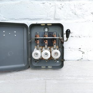 May include: An open, gray metal electrical fuse box with three white ceramic fuses. The box has a hinged lid and a black handle. The background is a white brick wall and a light gray wooden floor.